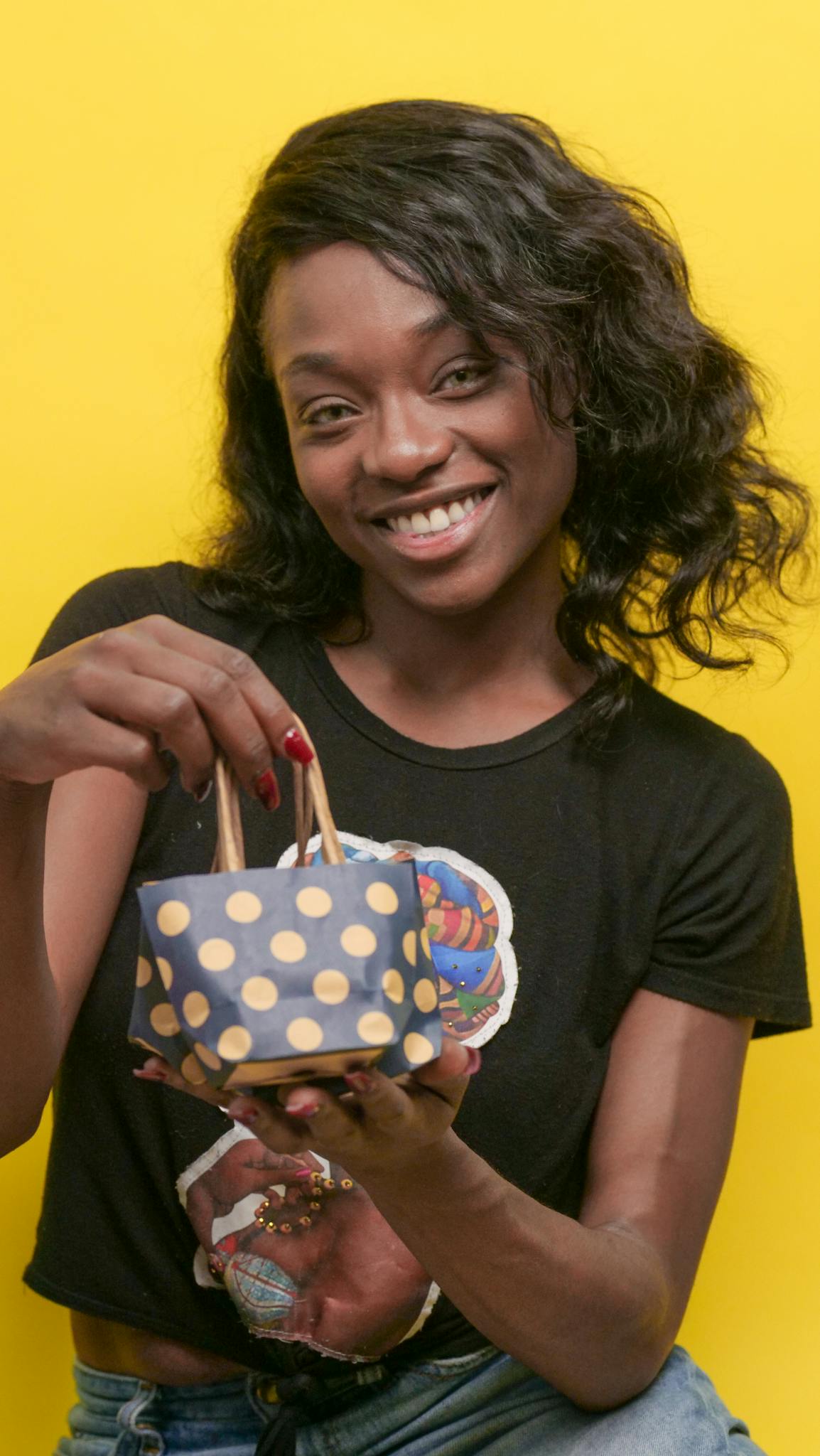 Cheerful woman holding a polka dot gift bag against a vibrant yellow background.